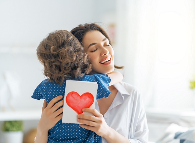 Happy mother's day! Child daughter is congratulating mom and giving her postcard. Mum and girl smiling and hugging.