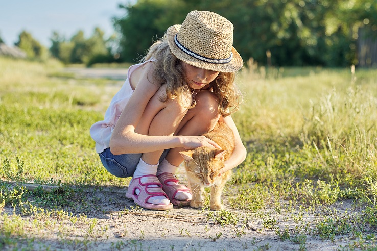 Girl child in hat on country road plays with red cat, sunny summer day rustic style