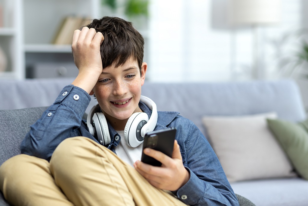 A smiling child of a teenage boy in headphones sitting on the sofa at home, resting and using the phone. Listens to music, watches videos. He chats, sits on social networks, plays online games