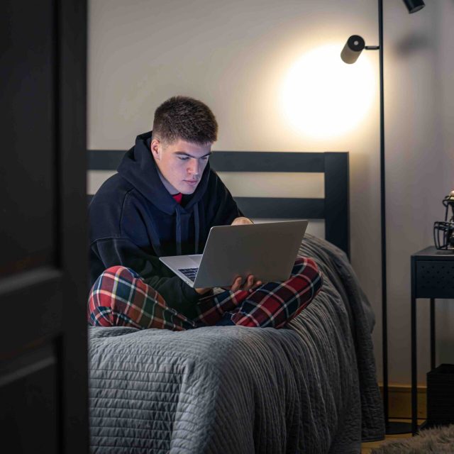 A teenager guy sits in a room on a bed and uses a laptop, a protective sports helmet on the nightstand, the concept of sports.