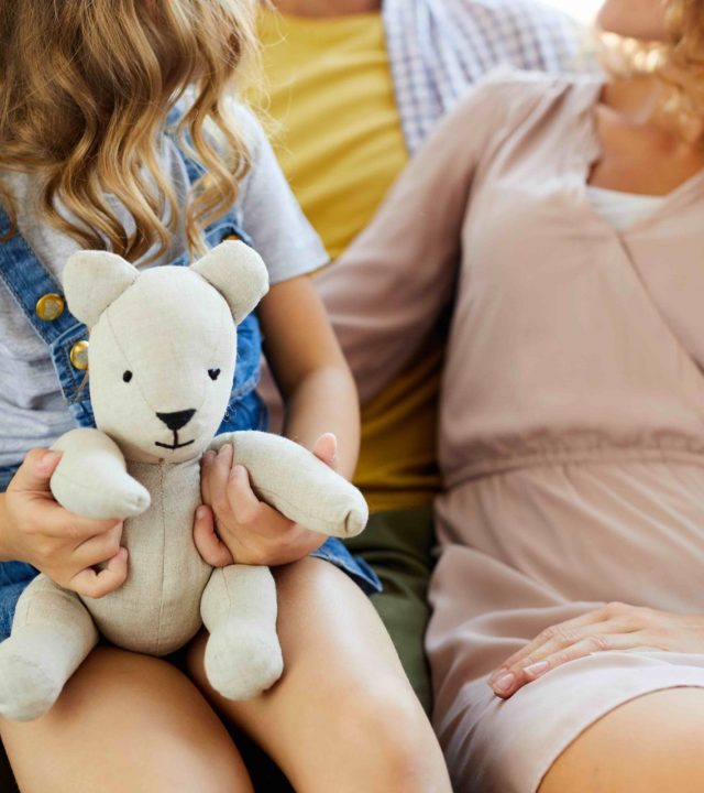 Little girl holding white teddybear with her mom and dad sitting near by