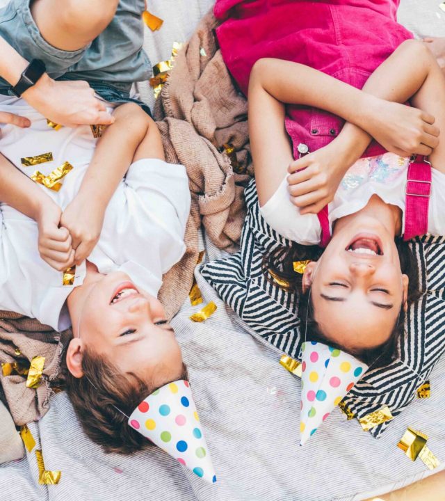 Little children laughing on plaid with confetti and air balloons at birthday celebration in cottage yard in summer.