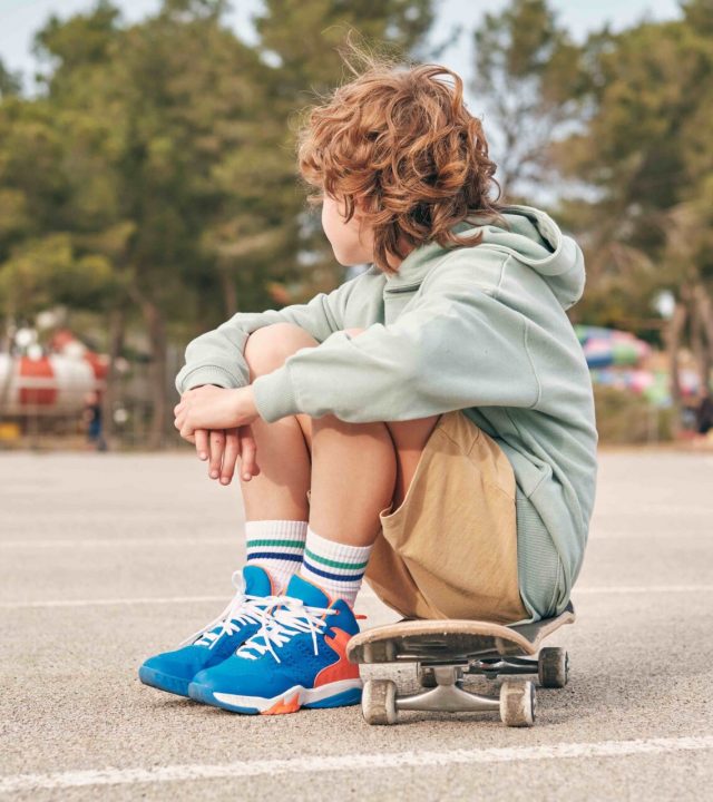 Side view of anonymous teen boy in cool outfit sitting on skateboard and embracing knees in street